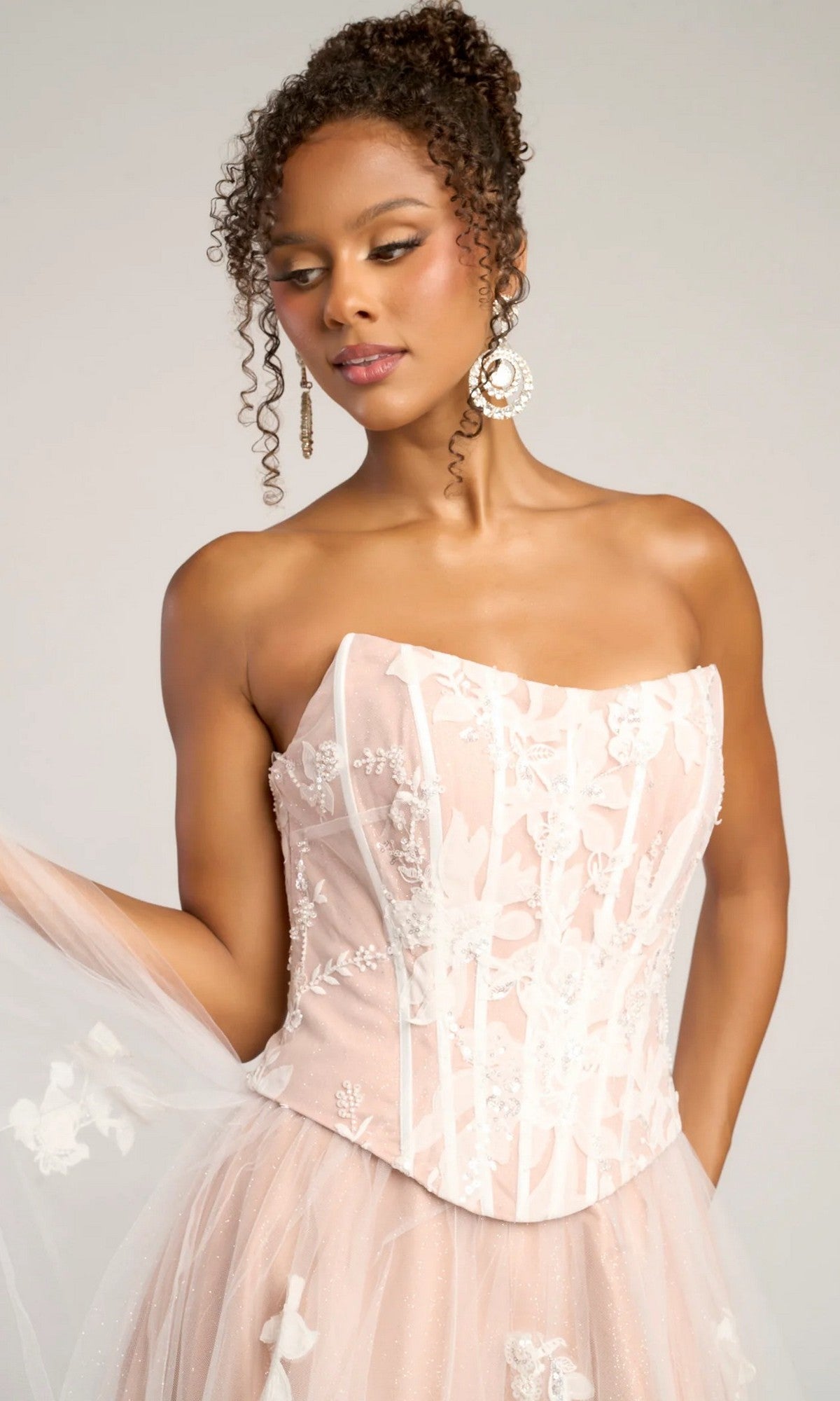 A woman wears the Portia and Scarlett PS26061 long formal dress—a strapless blush pink gown with a corset bodice, white floral embroidery, glitter tulle skirt—accessorized with large statement earrings.
